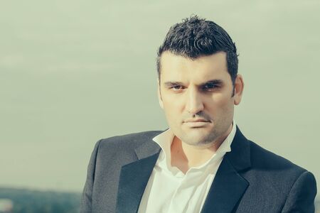 handsome male businessman with serious face in black jacket and white shirt outdoor on cloudy sky...