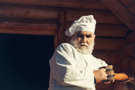 bearded man cook chef in uniform and hat with long beard holding wood cup with iron decoration sunny day outdoor on wooden backgroundの写真素材