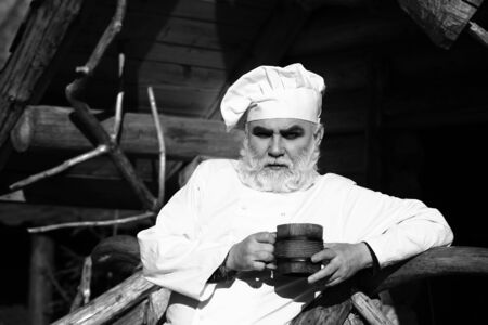bearded man cook chef in uniform and hat with long beard holding wood cup with iron decoration sunny day outdoor on wooden background, black and whiteの写真素材