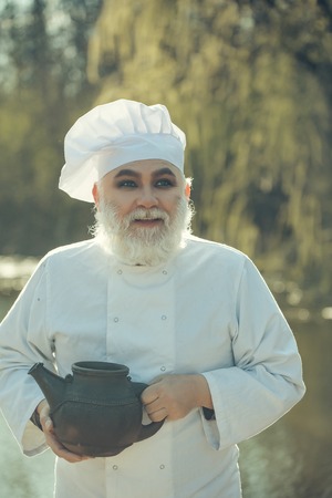 bearded man cook chef in uniform and hat with long beard on smiling face holding iron old tea kettle in blooming tree branches outdoor on natural backgroundの写真素材