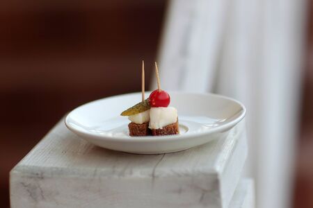 Canapes small snacks with lard rye bread pickled cucumber and cherry tomato on white plate on timber...