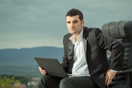 handsome male businessman with courageous face in black formal jacket and white shirt working on laptop sitting on leather office arm chair outdoor on cloudy sky backgroundの写真素材