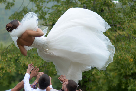 Men throw beautiful bride in white wedding dress and veil in air on green landscape backgroundの写真素材