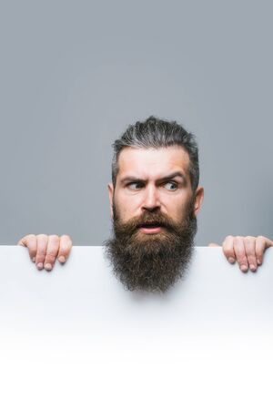 handsome bearded man with long lush beard and moustache on surprised face with white paper sheet in studio on grey background, copy spaceの写真素材
