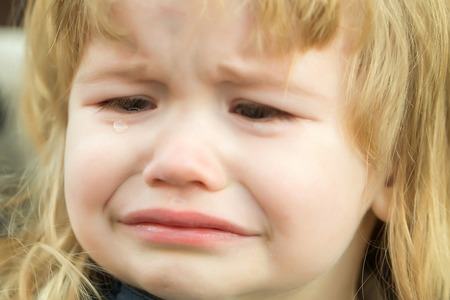 Little boy with cute emotional face and blond long hair cries with tears outdoor