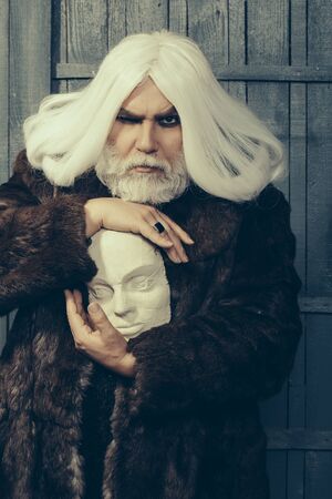 old druid bearded man with long beard on serious face and hair in fur coat holding white sculpture head in hands with ring on wooden backgroundの写真素材
