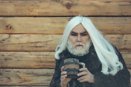 Druid old man with long grey hair and beard on serious face with wooden mug in hands outdoor on wood backgroundの写真素材