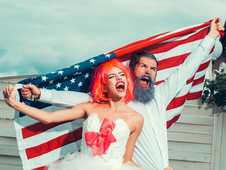 Happy young patriotic American couple with bright make up and hair celebrating Independence Day on 4th of July. American flag on the background, girl with the bright orange hairの写真素材