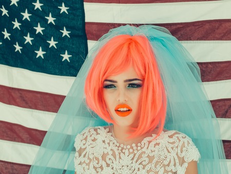 young patriotic pretty woman with red hair and national makeup in white dress and blue wedding veil on american flag background sunny weather celebrating independence day usaの写真素材