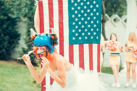 Ukraine, Kyiv - July 27, 2016: young patriotic sexy woman with pretty face and orange hair in soccer drink helmet with coca cola bottles on american flag background celebrating independence day usaのeditorial素材