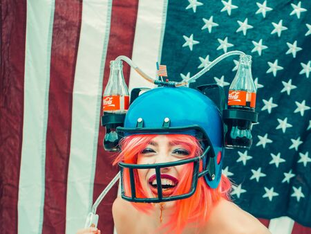 Ukraine, Kyiv - July 27, 2016: young patriotic sexy woman with pretty smiling face and orange hair in soccer drink helmet with coca cola on american flag background celebrating independence day usaのeditorial素材