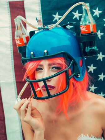 Ukraine, Kyiv - July 27, 2016: young patriotic sexy woman with pretty face and orange hair in soccer drink helmet with coca cola bottles on american flag background celebrating independence day usaのeditorial素材