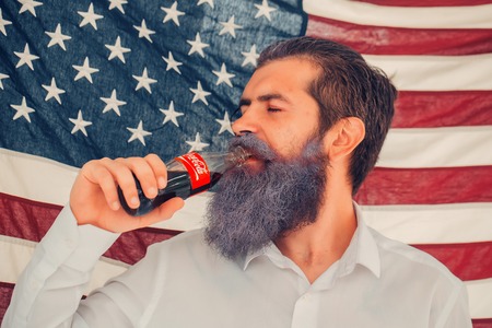Ukraine, Kyiv - July 27, 2016: young patriotic happy bearded man with long blue beard on american flag background drinking coca cola from bottle outdoor celebrating independence day usaのeditorial素材