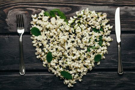 Fork and knife of stainless steel with heart wreath of white acacia blossoming flower petals green leaves on dark timber backgroundの写真素材
