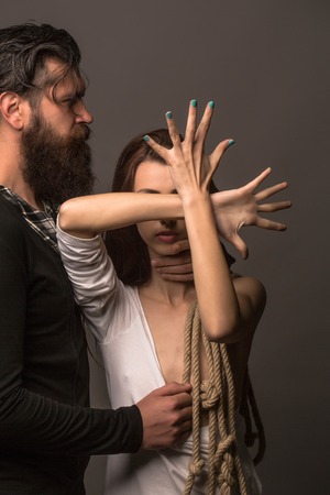 young couple of pretty naked woman with bare chest has rope on hands and handsome beard man with long beard touching female breast on grey backgroundの写真素材