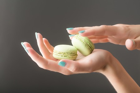 female hands with soft tender skin and blue manicure holding french biscuit of macaron or macaroon green color on grey backgroundの写真素材