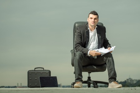 handsome male businessman in black formal jacket and white shirt with laptop and paper on leather arm chair outdoor with briefcase on cloudy sky backgroundの写真素材