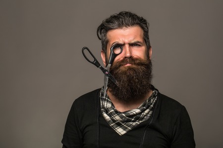 young handsome bearded man with long beard moustache and brunette hair holding hairdresser or barber scissors with emotional face in studio on grey backgroundの写真素材