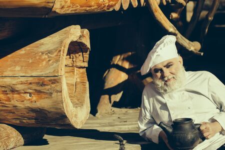 bearded man cook chef in uniform and hat with long beard on smiling face holding iron old tea kettle sunny day outdoor on wooden backgroundの写真素材