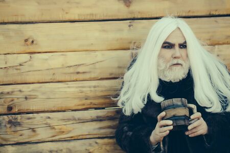 Druid old man with long grey hair and beard on serious face with wooden mug in hands outdoor on wood backgroundの写真素材