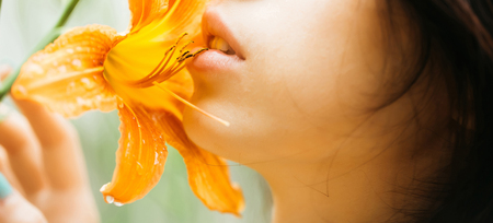 Sensual rosy lips on pretty woman face kiss bright orange lily flower blossoming on sunny dayの写真素材