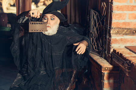 Old wizard with rusty coal iron in black costume and hat for Halloween on kitchen backgroundの写真素材