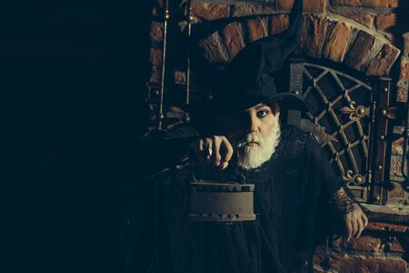Old wizard with rusty coal iron in black costume and hat for Halloween on kitchen backgroundの写真素材