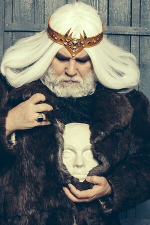old druid bearded man with long beard on serious face and hair in fur coat and crown holding white sculpture head in hands with ringの写真素材