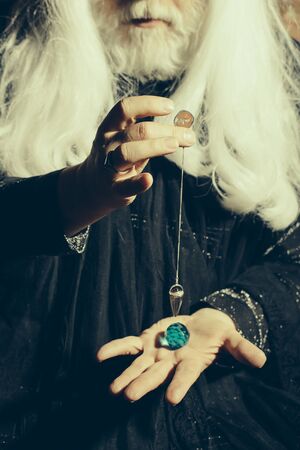 Magic pendulum in hands of old wizard with long silver hair and beard in black costume for Halloweenの写真素材
