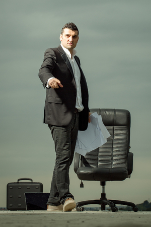 handsome tired male businessman in black formal jacket and white shirt with laptop and paper near leather arm chair outdoor with briefcase on cloudy sky backgroundの写真素材