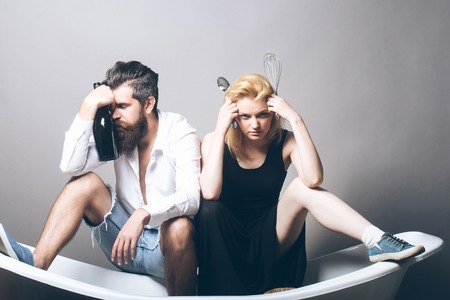 young family couple of blonde pretty girl with kitchen utensils and bearded man with long beard holding wine bottle sitting on bathtub. household, everyday life and routineの写真素材