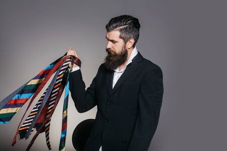 Young man with handsome serious face and stylish beard holding many different ties in his hand posing on gray background studioの写真素材