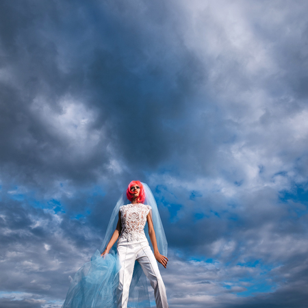 young pretty woman with orange or pink hair and bright makeup on emotional face in white wedding suit and blue bride veil on natural cloudy sky background outdoorの写真素材