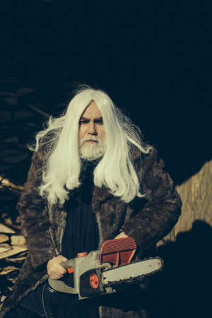 Old man druid with long silver hair and beard in fur coat holding big chainsaw sunny day outdoor