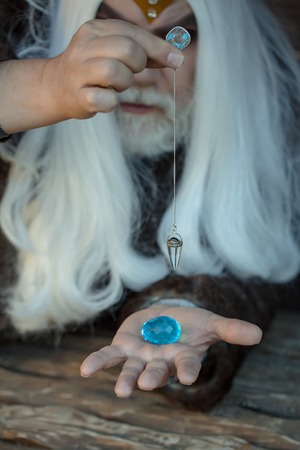 Old bearded man wizard with long white hair and beard holding blue gem stone and silver pendant for...