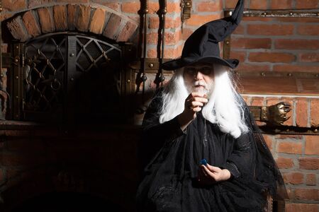 Old wizard with pendulum in black costume and hat for Halloween on kitchen background