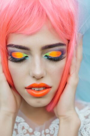 Young girl with attractive face with closed eyes bright professional fashion eye shadow makeup orange lips and pink wig holding her hand by handsの写真素材