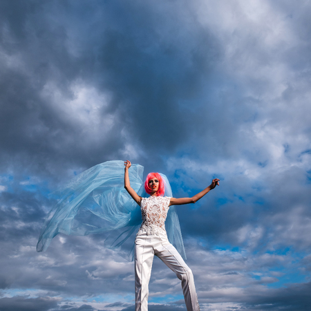 young pretty woman with orange or pink hair and bright makeup on emotional face in white wedding suit and blue bride veil on natural cloudy sky background outdoorの写真素材