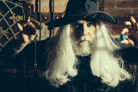 Old man wizard with long gray hair beard in black costume and hat for Halloween holding blue gem stone and silver pendant for hypnosis on wooden backgroundの写真素材