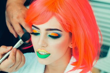 Young girl model with pretty sexy face green yellow lips orange hair and makeup artist applying colorful eye shadow by brushの写真素材