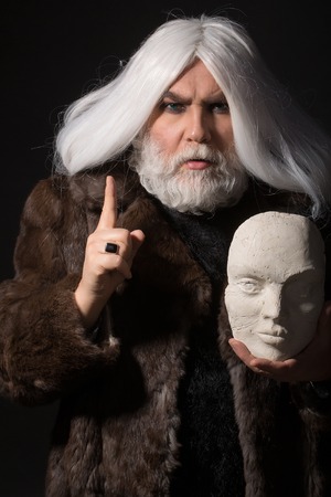 old druid bearded man with long beard on serious face and hair in fur coat holding white sculpture head in hands with ring on dark backgroundの写真素材