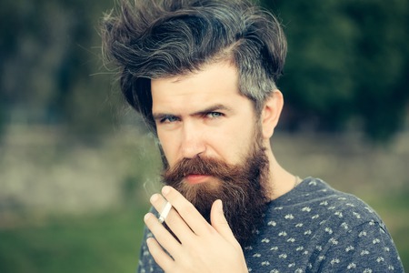 handsome sexy bearded young man hipster with long beard and mustache has stylish hair on serious hairy face smoking cigarette outdoor on green natural background, closeupの写真素材