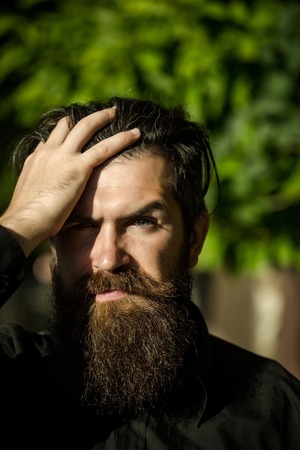 handsome sexy bearded young man hipster with long beard and mustache has stylish hair on serious hairy face sunny day outdoor on green natural background, closeupの写真素材