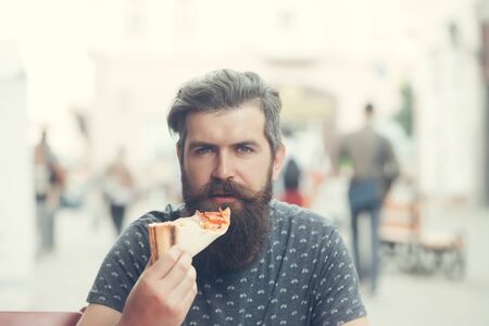 handsome sexy bearded young man hipster with long beard and mustache on serious hairy face eating pizza on street outdoorの写真素材