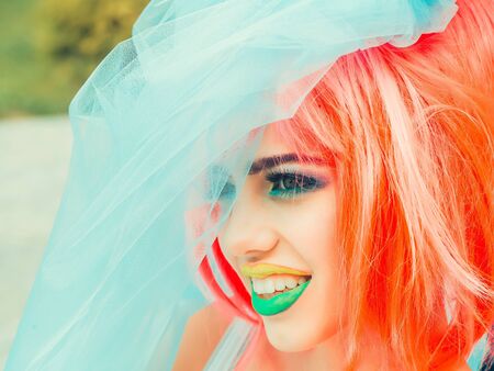 Young girl with pretty face bright professional makeup colorful eyeshadow yellow and green lips and orange hair wig wearing blue veilの写真素材