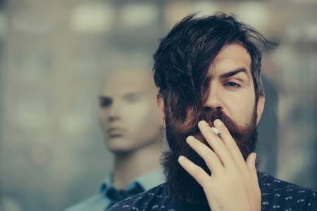 handsome bearded young man hipster with long beard and mustache has stylish hair smoking cigarette near shop glass showcase with dummy in shirt and tieの写真素材