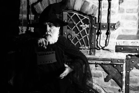 Old wizard with rusty coal iron in costume and hat for Halloween black and white on kitchen backgroundの写真素材