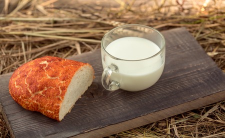 Freshly baked loaf of wheat bread and cut slice on wooden board on straw or hay background with glass cup of milkの写真素材