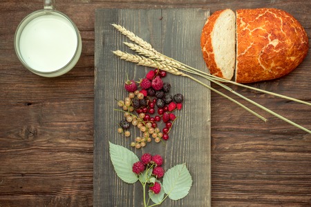 Freshly baked loaf of wheat bread and cut slice on wooden board with spikelet red and white currant strawberry blackberry raspberry and glass cup of milkの写真素材