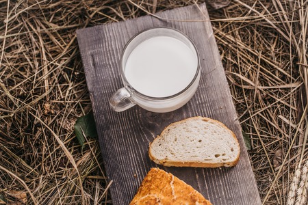 Freshly baked loaf of wheat bread and cut slice on wooden board on straw or hay background with glass cup of milkの写真素材